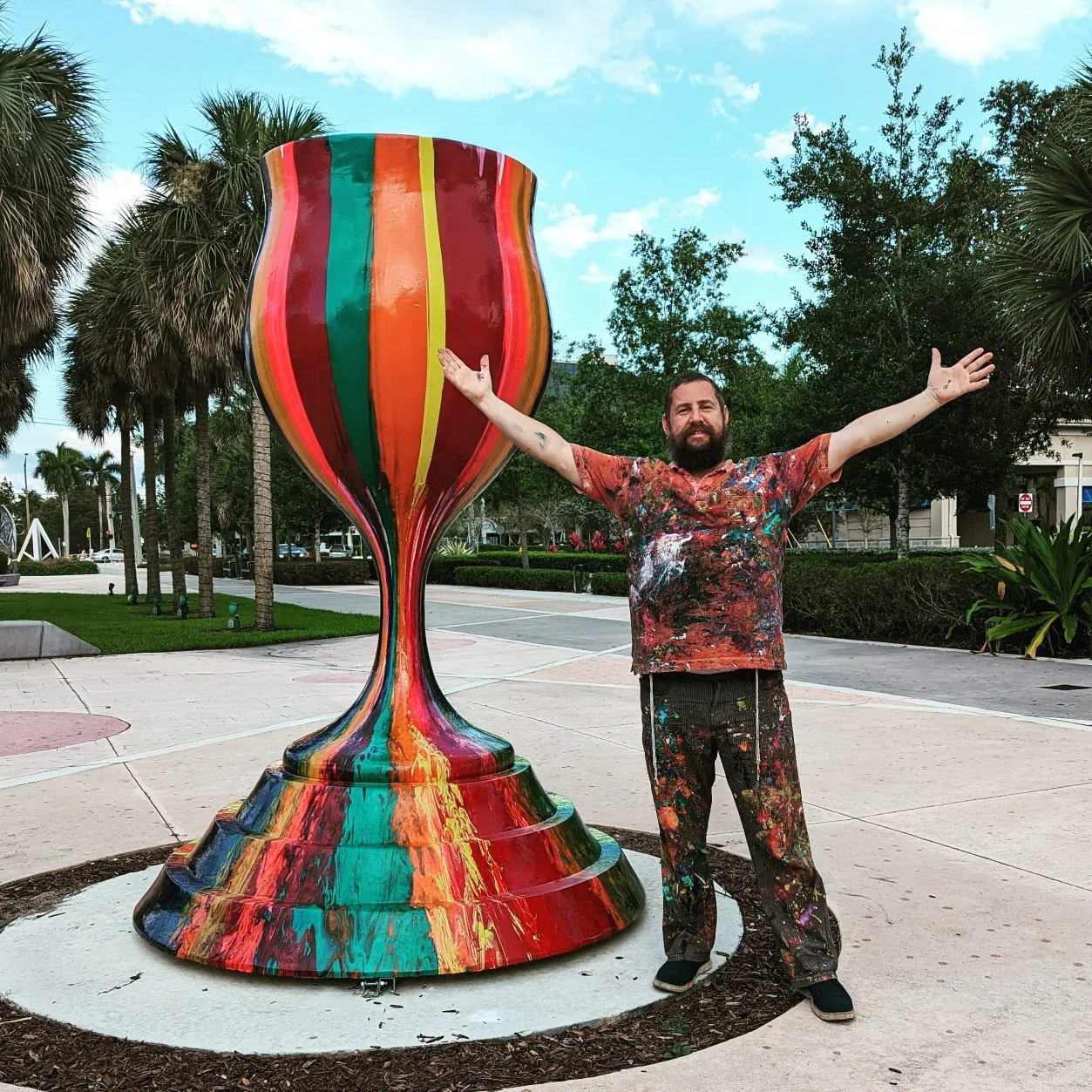 Yitzchok Moully standing next to his Tradition With a Twist public art menorah — large colorful Jewish sculpture, outdoor installation with palm trees