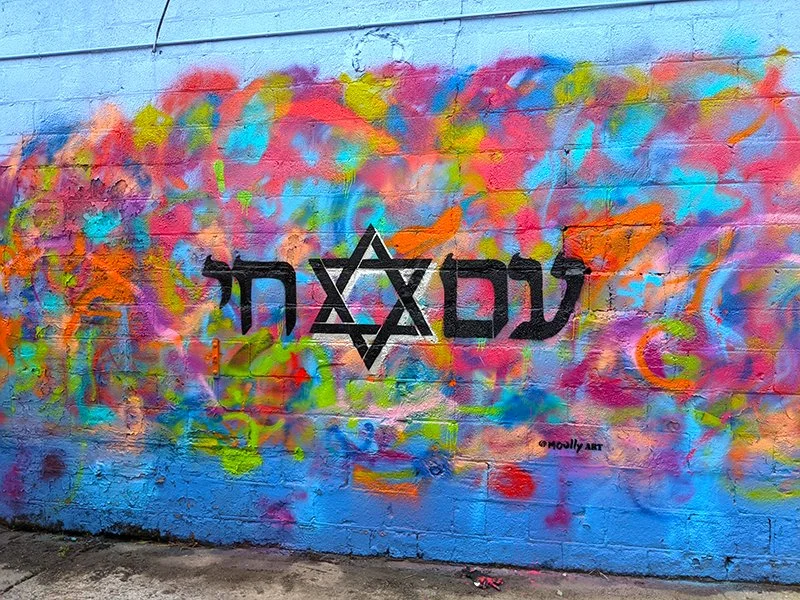 Graffiti wall with multicolored abstract paint background and black spray-painted Hebrew text centered over a Star of David.