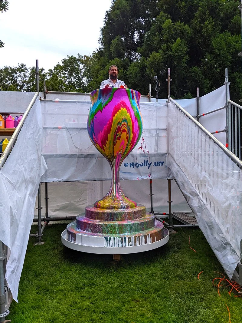 A man standing inside a large, colorful, hand-painted goblet sculpture on a platform in an outdoor setting with trees and scaffolding around.