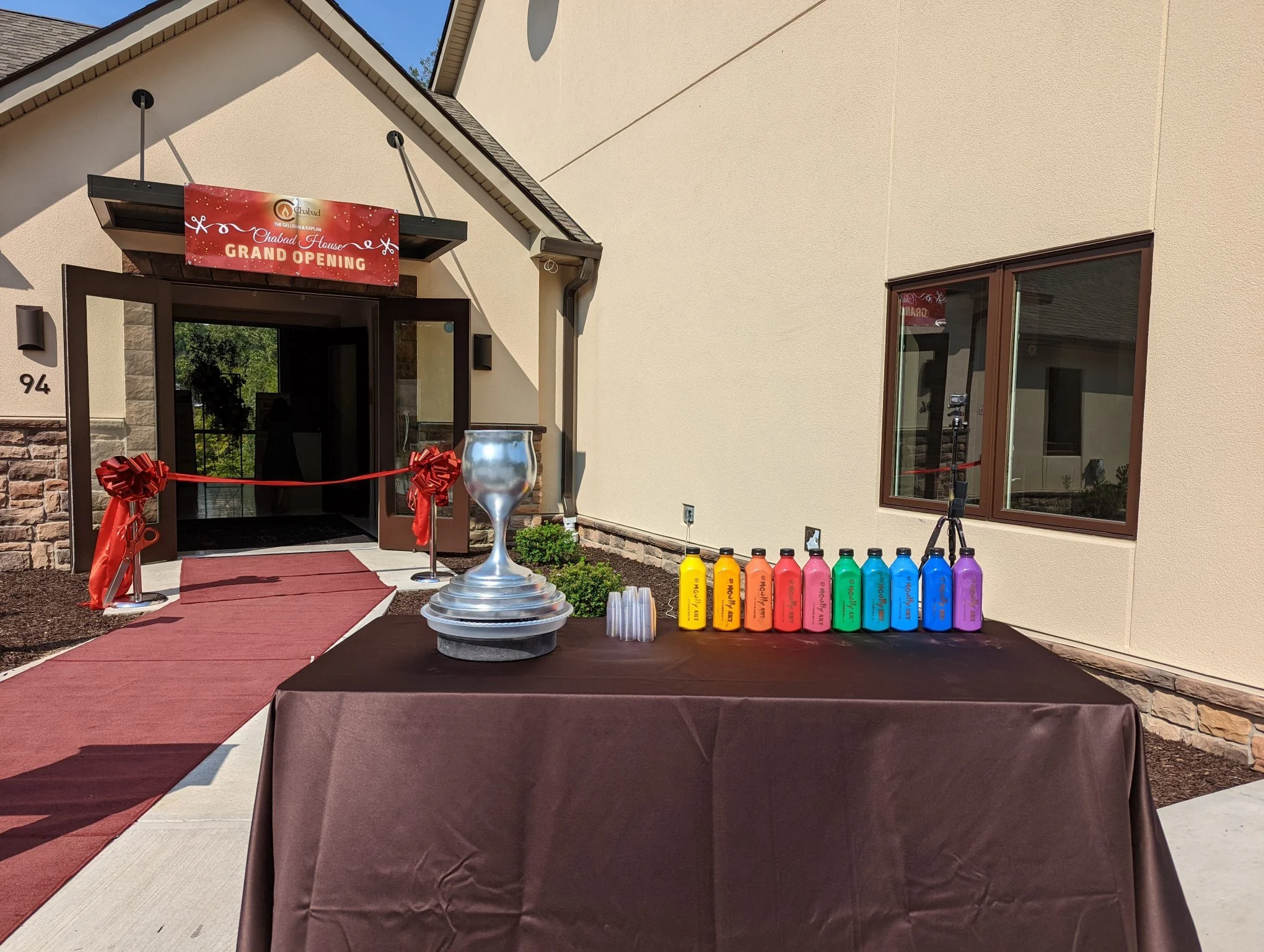 Entrance to a building decorated for a grand opening event, with a red banner above the door that reads "Chabad House Grand Opening," red ribbon, red carpet, colorful bottles of paint or drinks on a table, and a large silver goblet on a table in the 