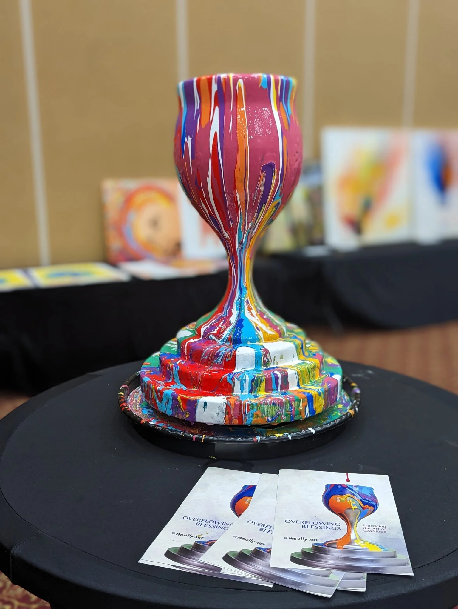 Colorful painting of a vase with multicolored drips of paint running down its surface, placed on a black table with brochures titled 'Overflowing Blessings' featuring an image of the vase painting and the subtitle 'Practicing the Art of Gratitude'.