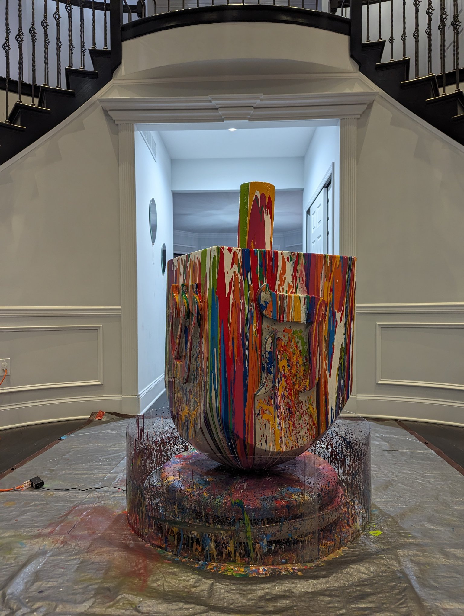 Large colorful Jewish art sculpture by Yitzchok Moully being painted in studio — vibrant drip-paint installation in progress, gallery with plastic floor covering