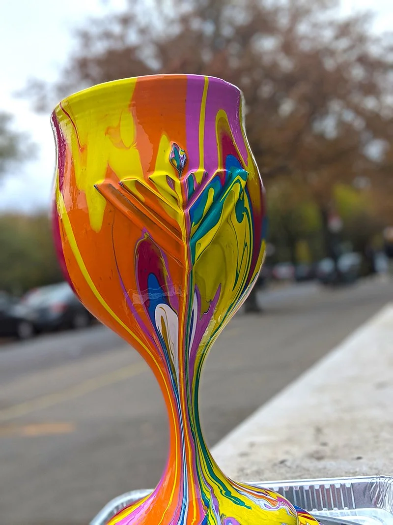 A colorful, abstract marble of paint with vibrant swirls of yellow, orange, purple, blue, green, and white, set outdoors with a blurred background of trees and a parking lot.