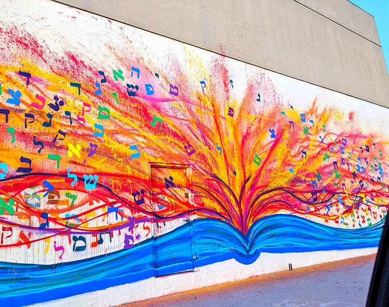 Colorful mural of a book with pages opening, from which vibrant musical notes and abstract shapes are flowing outward in bright colors like red, yellow, pink, green, and blue.
