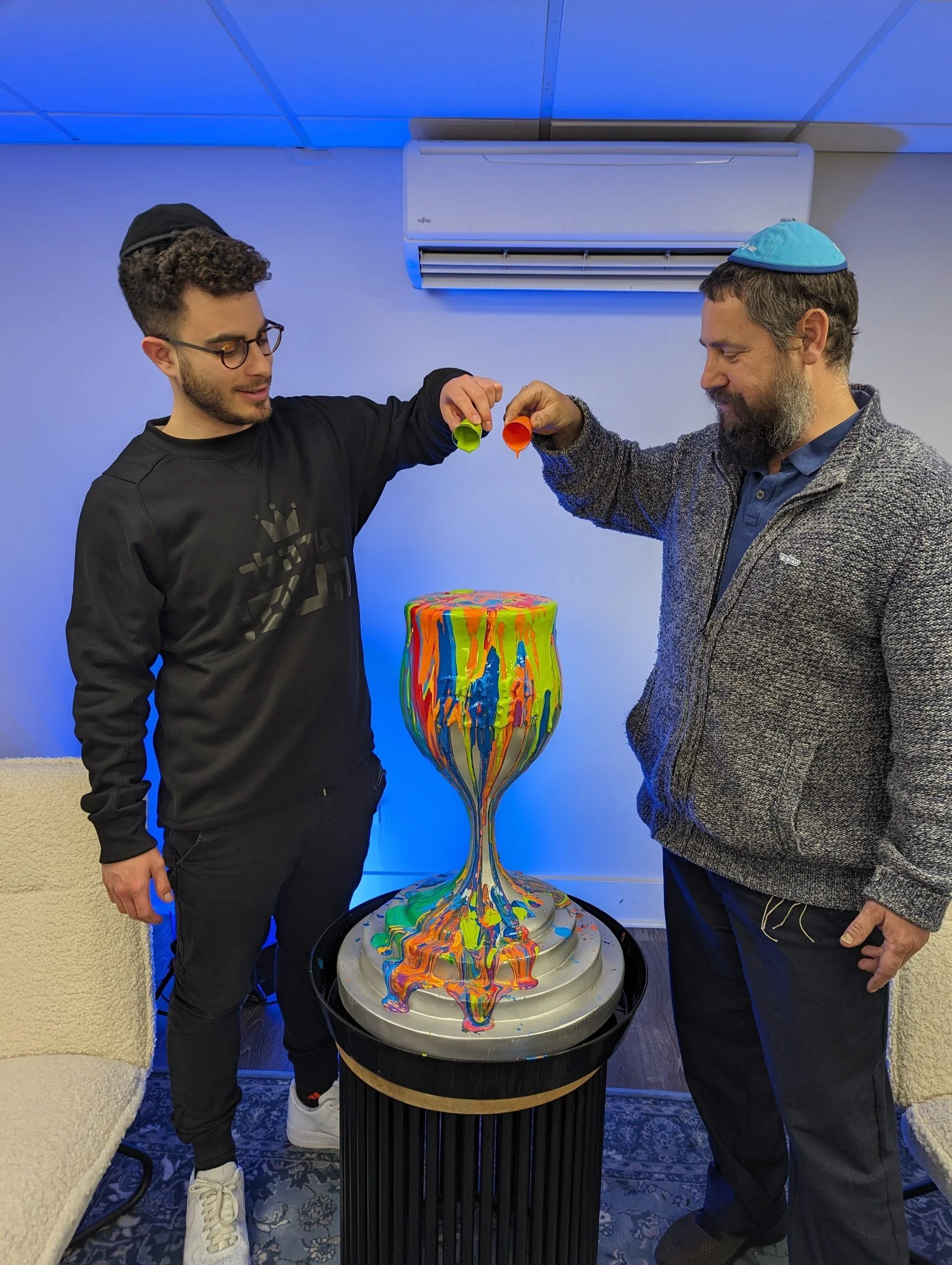 Jewish men participating in paint-pour activity at Yitzchok Moully community art workshop — kippah-wearing participants creating colorful canvas together