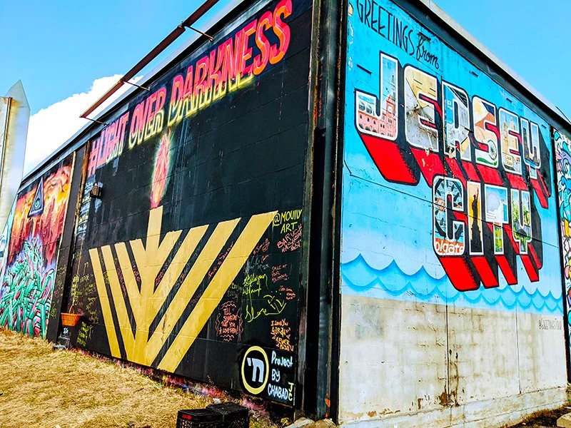 Colorful mural on a large outdoor wall with graffiti, featuring a welcoming message from Jersey City and various artistic designs, including a beach scene with waves.