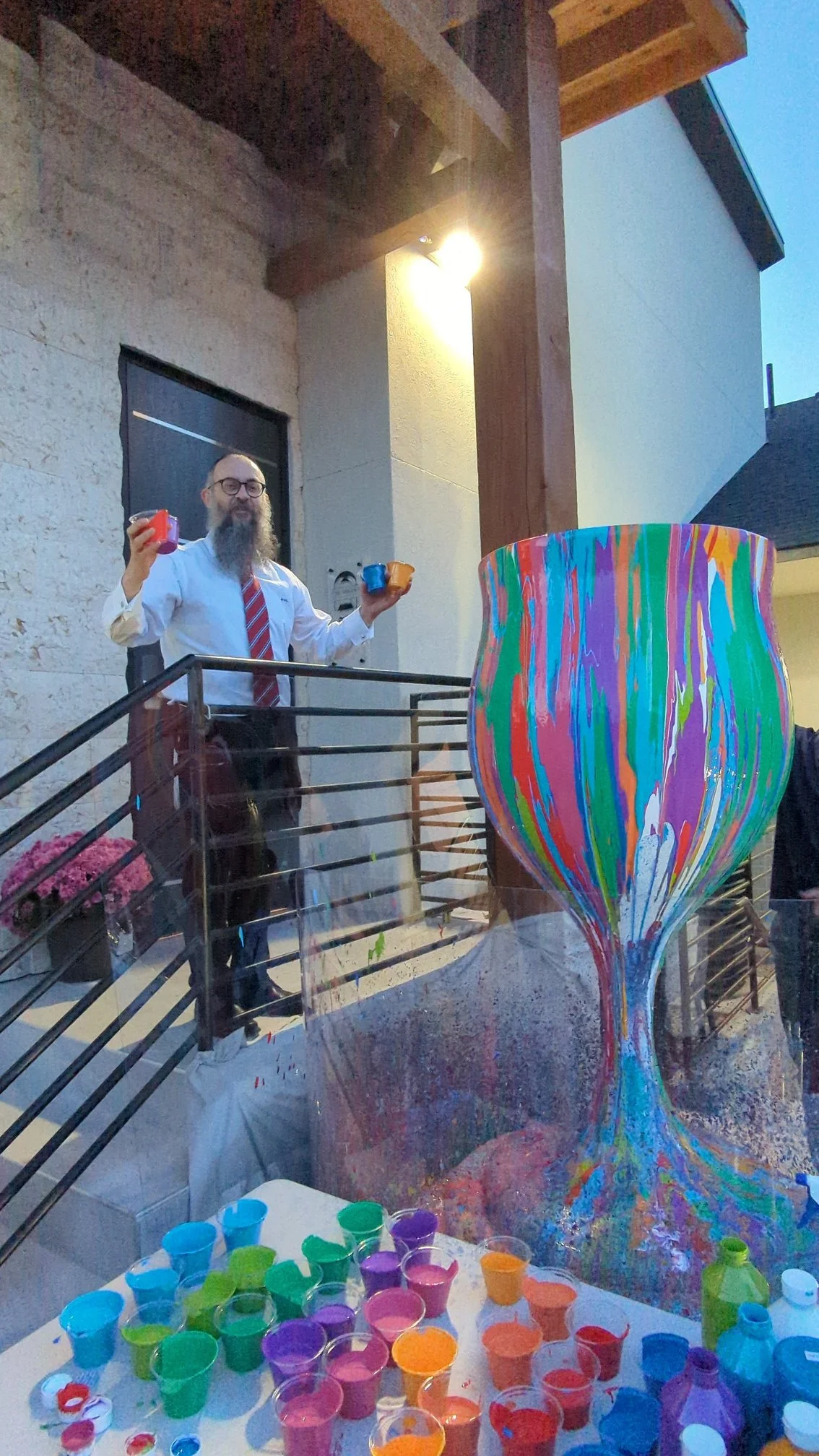 Yitzchok Moully with Overflowing Blessings kiddush cup sculpture at outdoor dusk Jewish art event — paint cups and colorful vase on balcony table