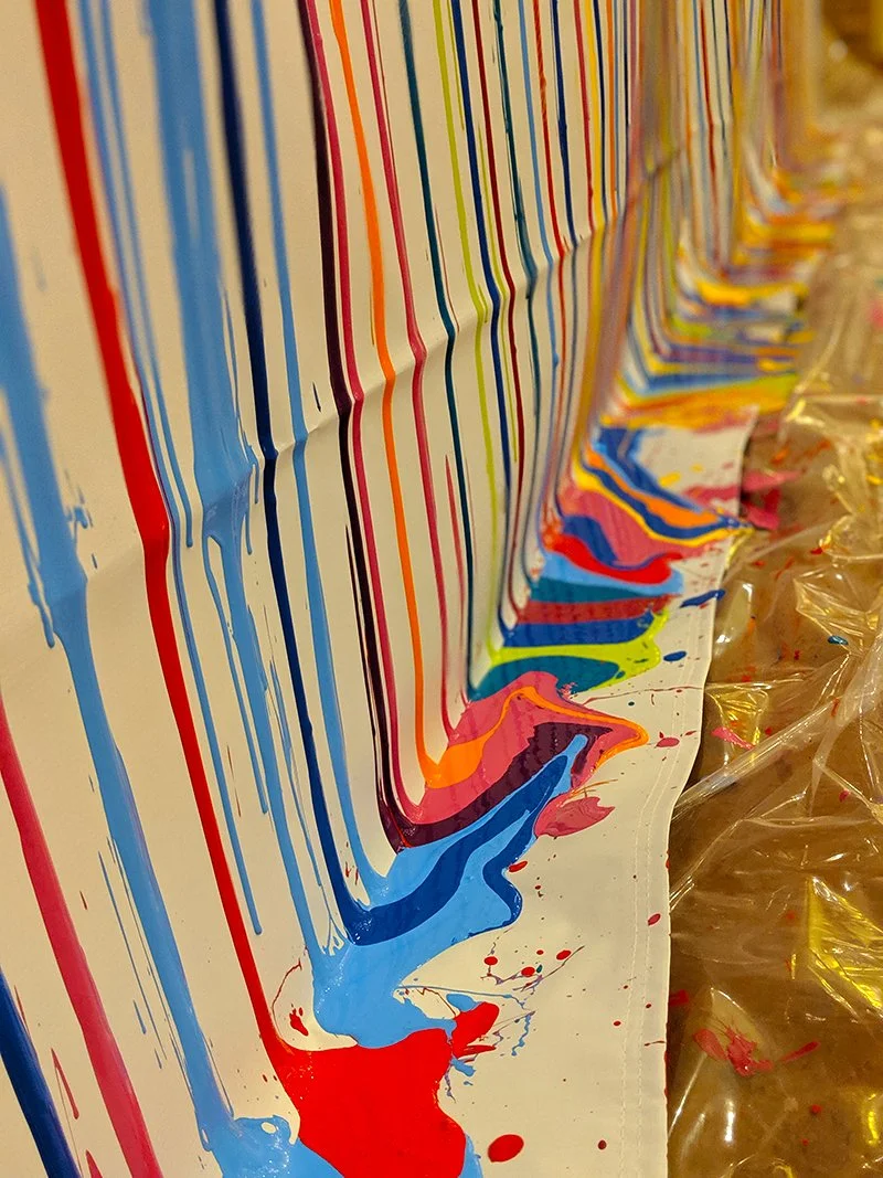 Close-up of a colorful abstract painting with vertical streaks of blue, red, orange, pink, green, and yellow paint on a white canvas, with some paint dripping and pooling at the bottom.