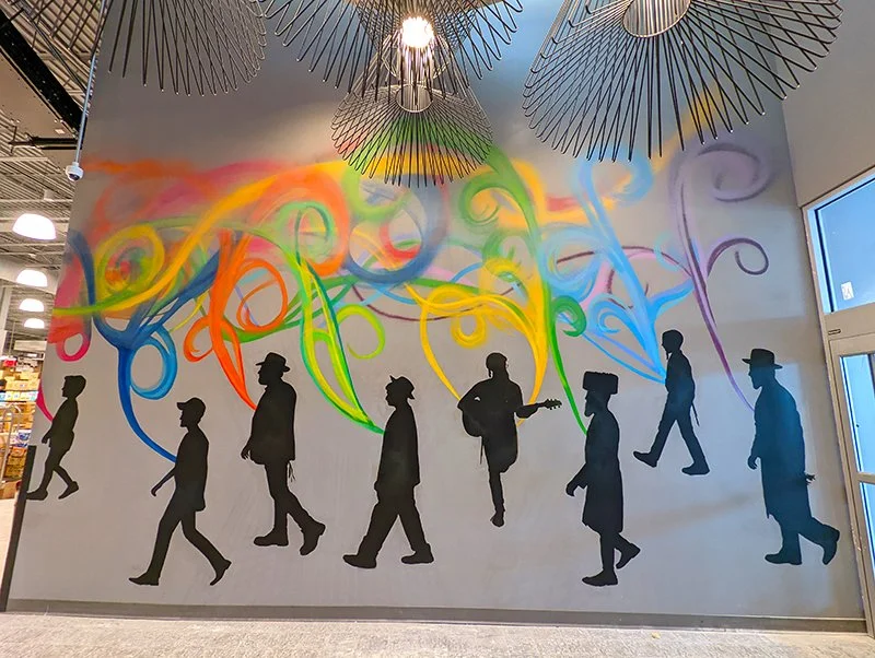 Silhouettes of people walking in a line with colorful, swirling spray paint above them on a wall, inside a building with modern light fixtures and large window.