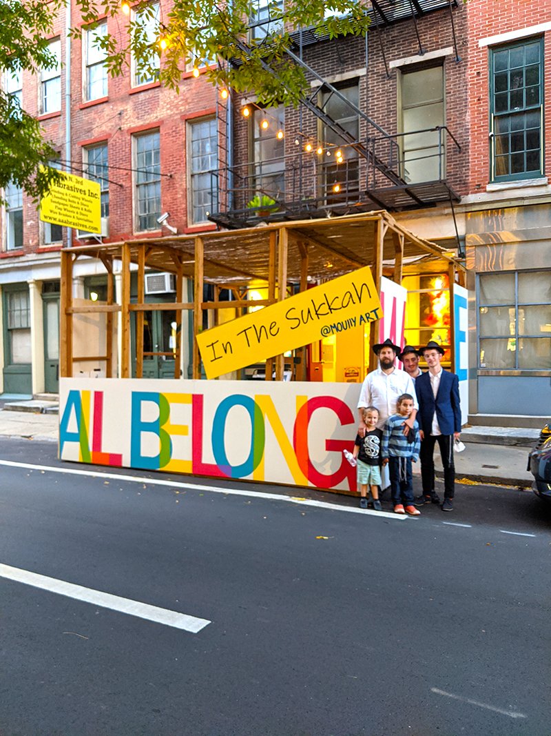 A colorful outdoor art installation on a city street with a large sign reading "ALBONG" in multicolored letters, a wooden structure, and a group of five people including children and adults posing in front of it. The background shows brick buildings 