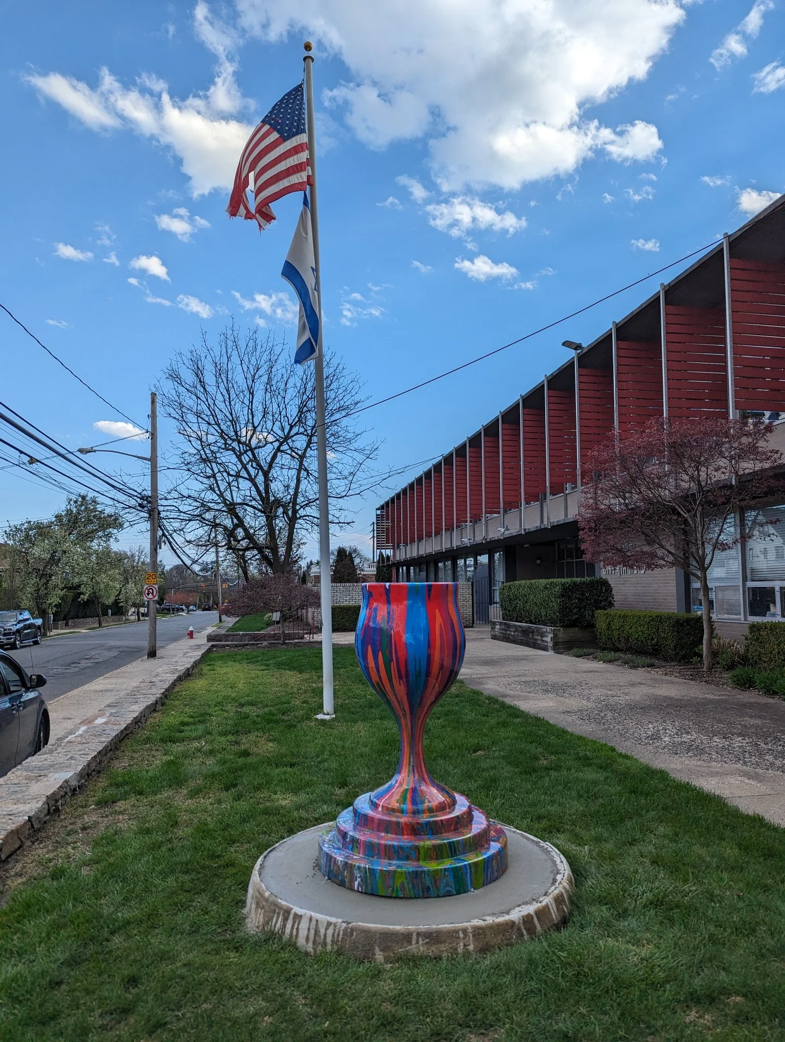 Colorful painted vase sculpture placed on a concrete platform in a grassy area, with flags, trees, and a building in the background under a blue sky with scattered clouds.