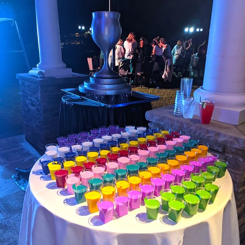 Colorful cups with mixed drinks arranged on a table at an outdoor event, with a large trophy in the background.