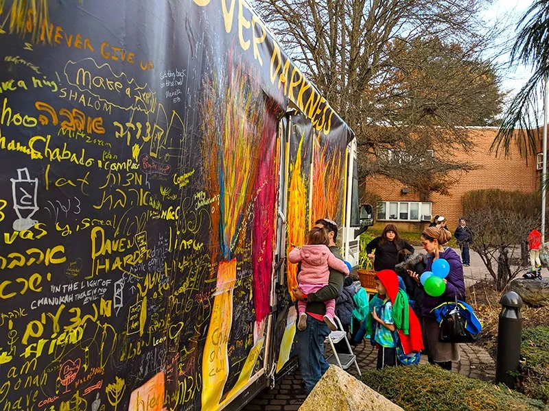 People gathering around a colorful truck with yellow and red paint, writing, and splash designs on it in an outdoor urban setting.