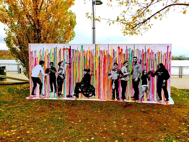 Colorful outdoor art installation featuring cutout silhouettes of people in a park with autumn trees and a river in the background.