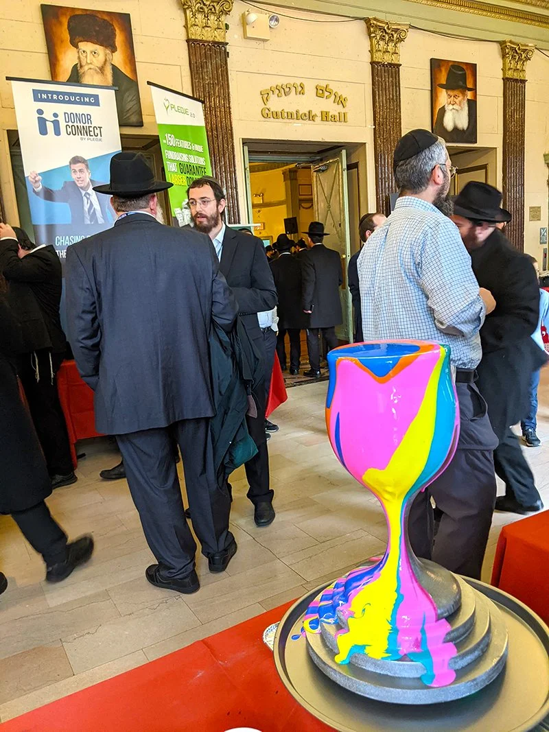 People gathered inside a synagogue, with some wearing traditional Jewish attire, near a colorful hourglass sculpture on a table. Portraits of rabbis are mounted on the wall, and signs for donor and event organizations are displayed.
