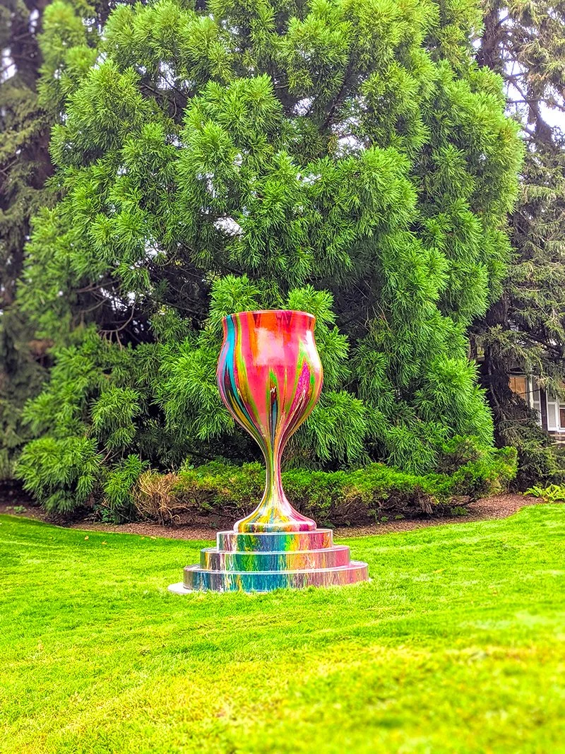 Colorful, abstract sculpture resembling a large, decorative goblet or vase, placed on a multi-tiered platform in a lush green garden with tall trees.