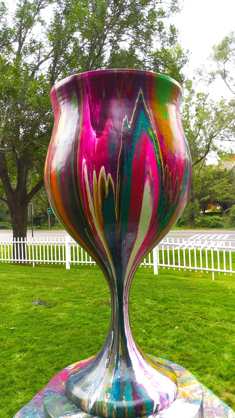 Colorful, abstract painted vase standing outdoors on green grass with trees and a white picket fence in the background.