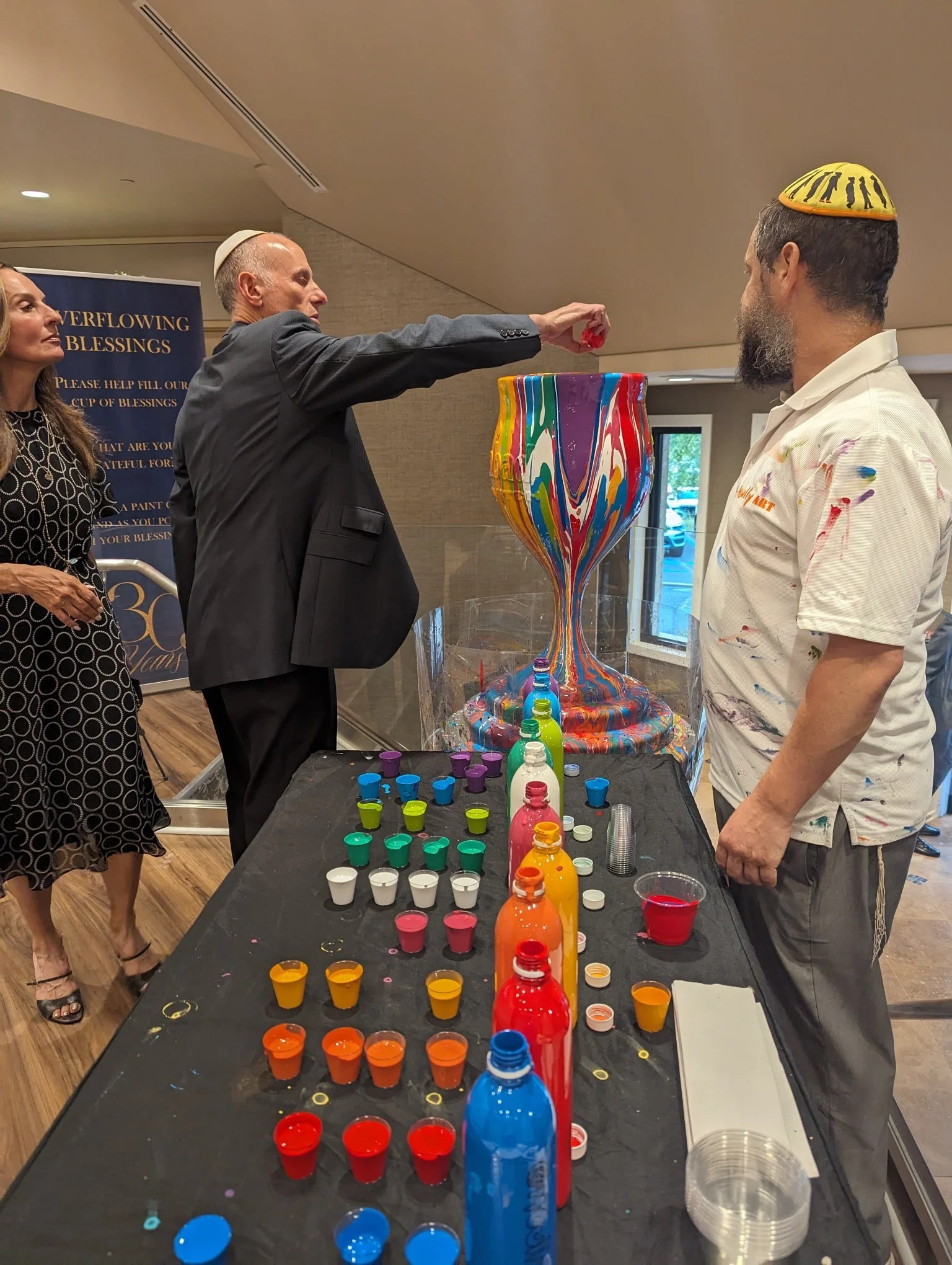 Hanukkah dreidel spinning event with Overflowing Blessings drip-paint menorah by Yitzchok Moully — festive Jewish community celebration with paint bottles
