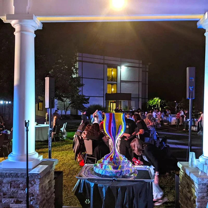 Colorful glass art sculpture on a table at an outdoor evening event, with people sitting and socializing in the background.
