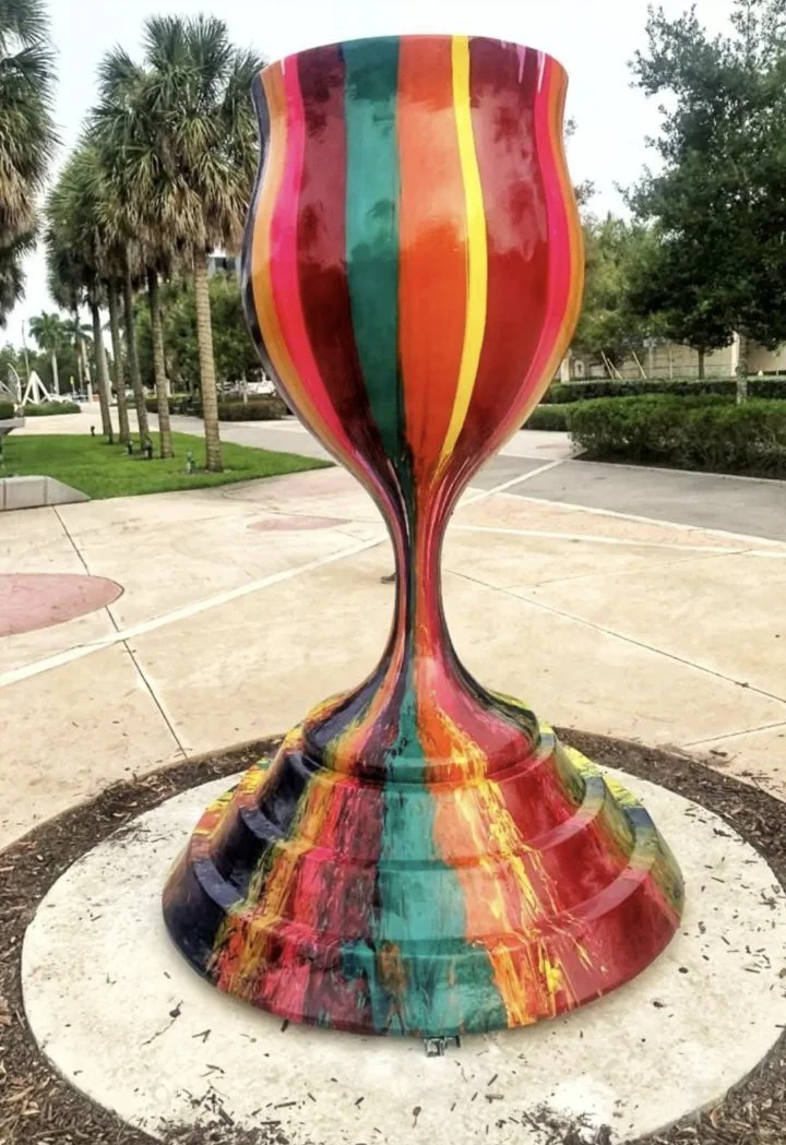 Colorful, abstract sculpture resembling a large, stylized goblet or trophy, painted with vertical stripes in red, yellow, green, blue, and orange, located outdoors on a concrete pad surrounded by trees and shrubs.