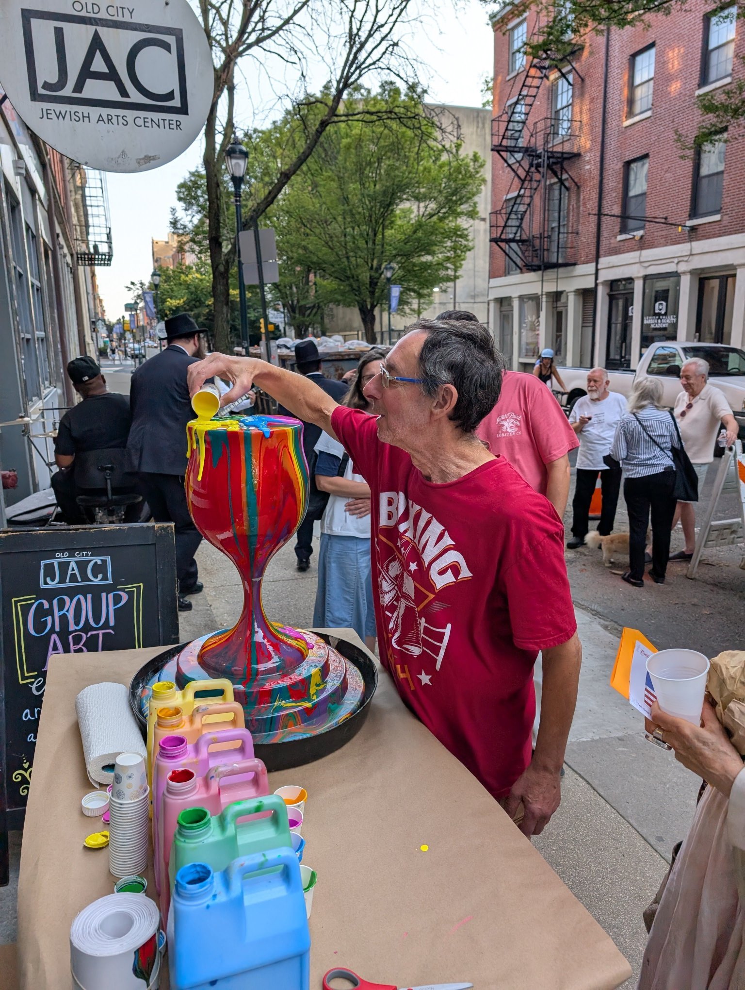 A man in a red shirt pouring bright yellow paint into a large, rainbow-colored ceramic vase at an outdoor art event on a city street. There are people in the background, some icons and signs, and a chalkboard that reads 'JAC Group Art'.