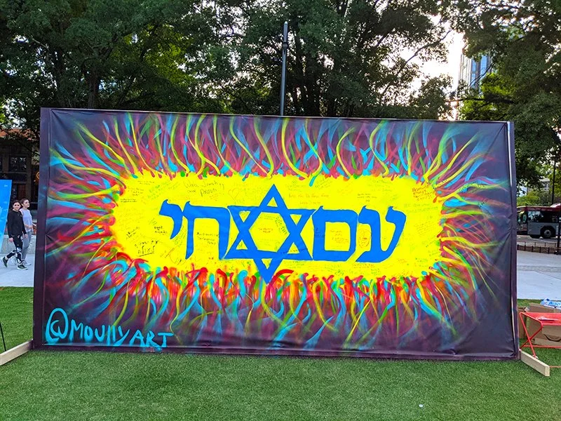 A colorful graffiti mural with Hebrew text, a Star of David, and the artist's handle @MOUNYART, displayed outdoors on a large banner.