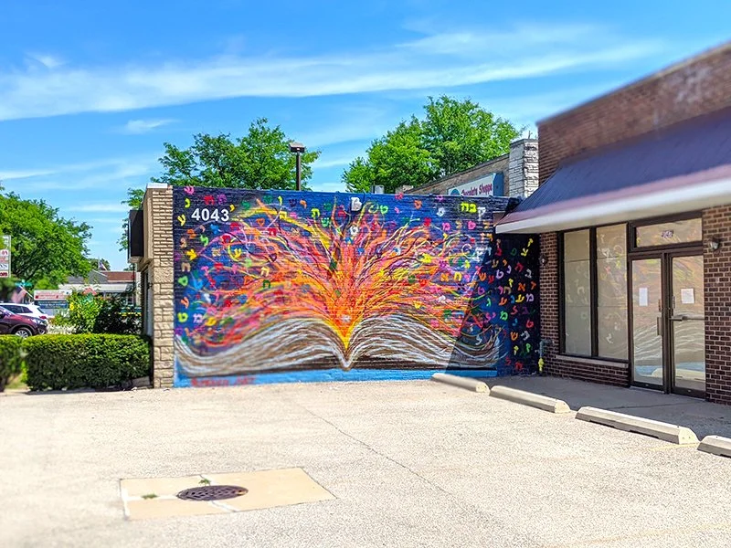 Colorful mural of an open book with pages turning into a vibrant explosion of multicolored flames and abstract shapes, painted on a brick wall outside a building.
