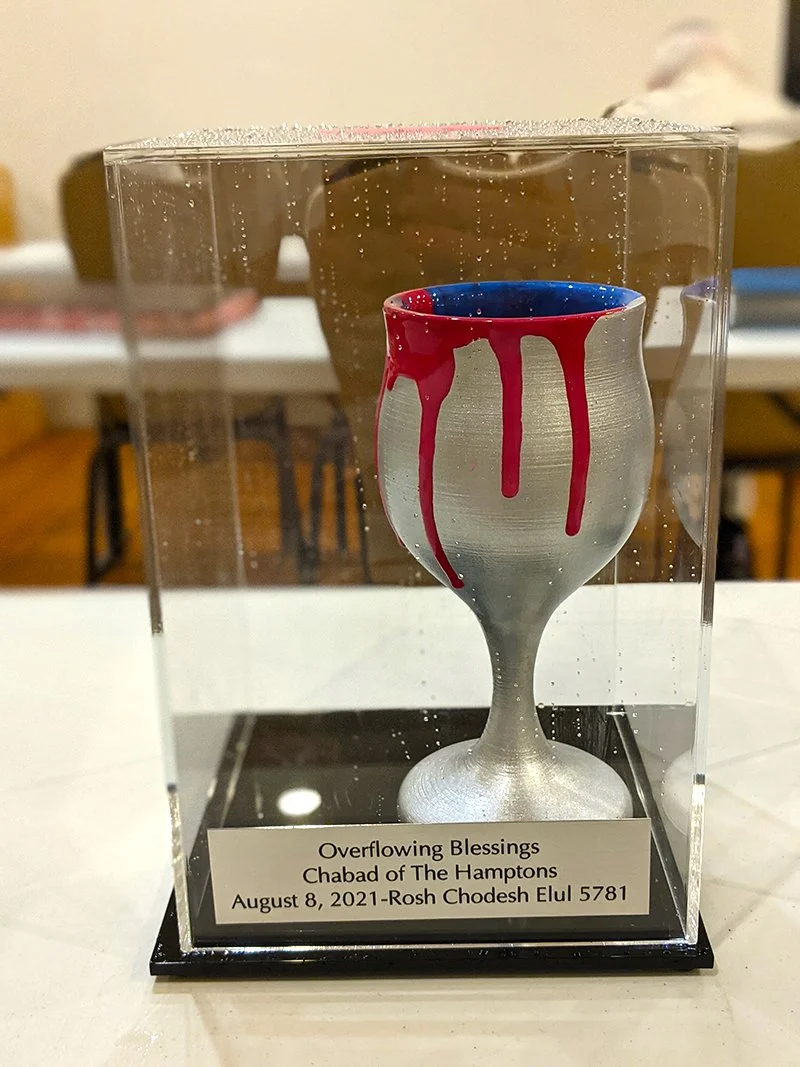 A decorative goblet with red and blue paint dripping from the top, encased in a clear display box, with a plaque reading "Overflowing Blessings, Chabad of The Hamptons, August 8, 2021, Rosh Chodesh Elul 5781."