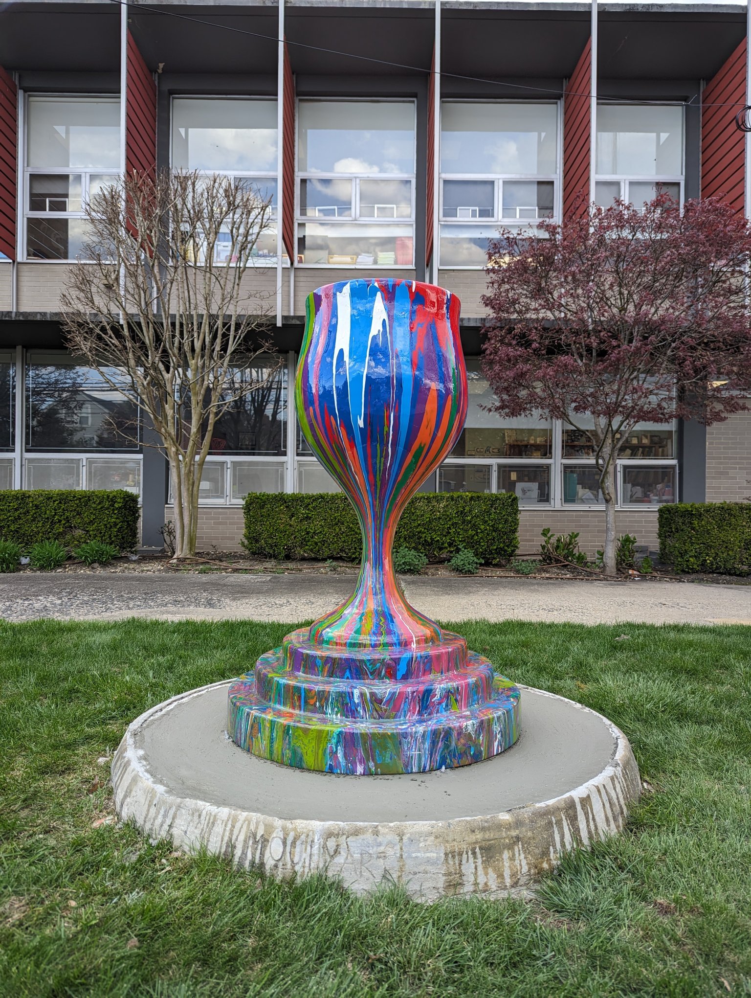 Overflowing Blessings outdoor kiddush cup sculpture by Yitzchok Moully — rainbow drip-paint Jewish art installation on circular base in manicured lawn, Jewish community center