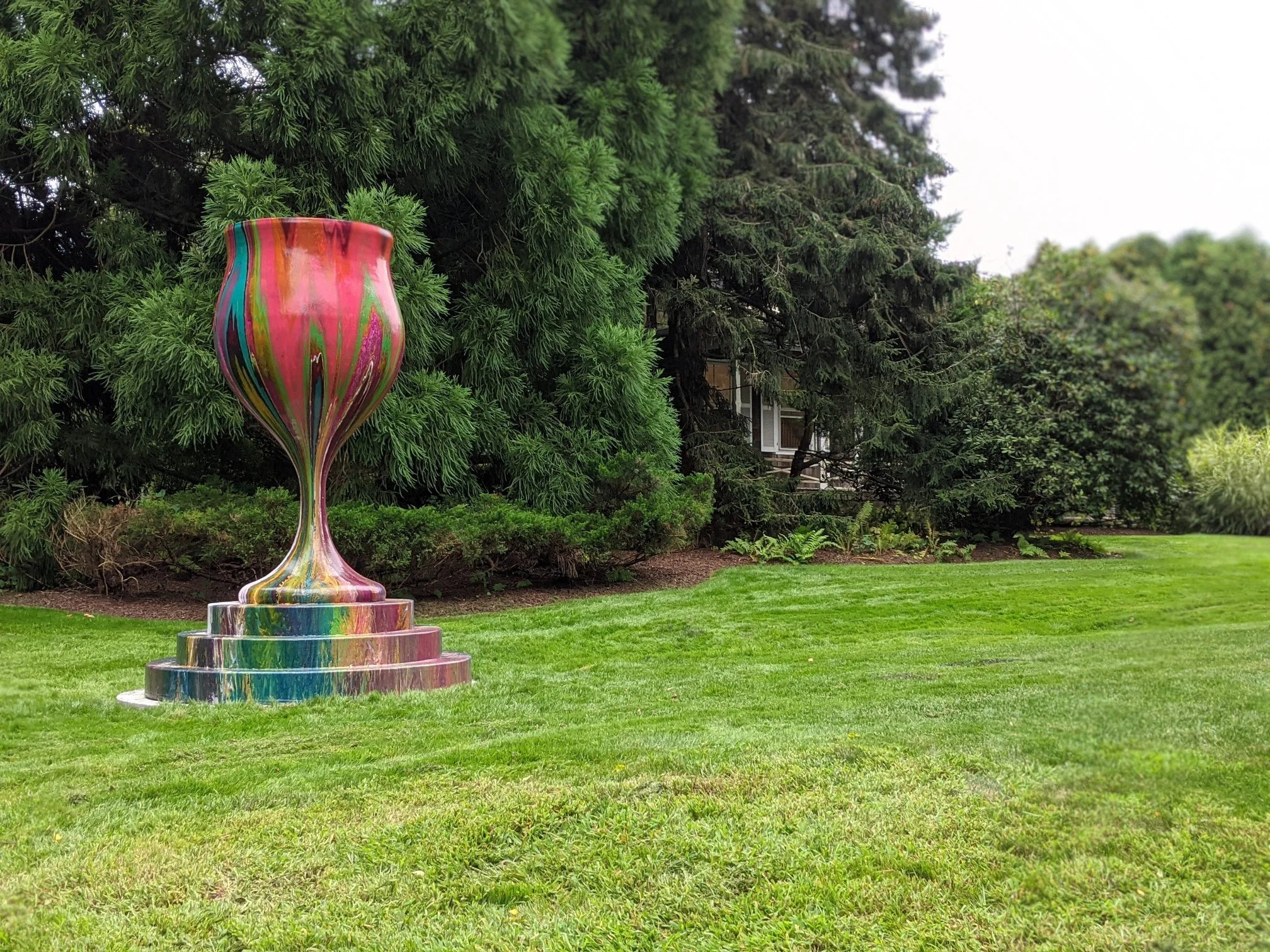 A colorful, marbled sculpture of a chalice or goblet on a tiered base outdoors in a green garden with trees and grass.