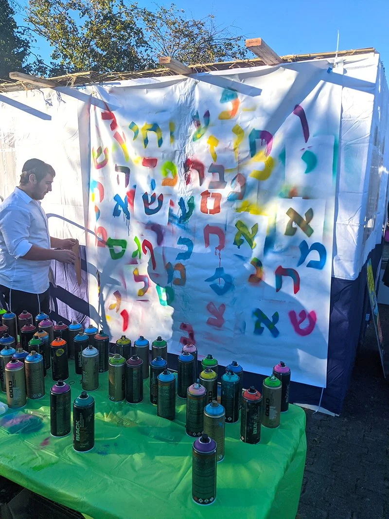 Colorful spray paint cans on a green table in front of a white wall with Hebrew letters spray painted in various colors.