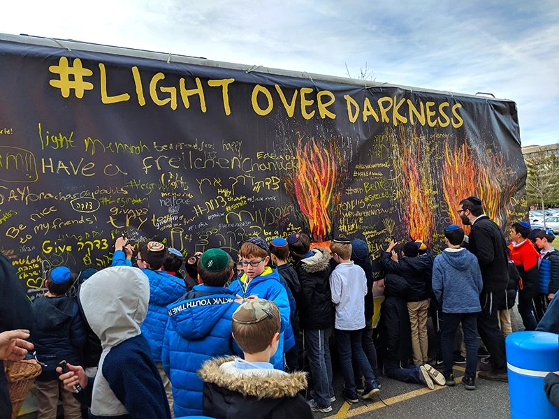 A group of children and adults gathered in front of a large black wall with yellow writings and drawings of flames, participating in an outdoor event with a banner reading '#LIGHT OVER DARKNESS' at the top.
