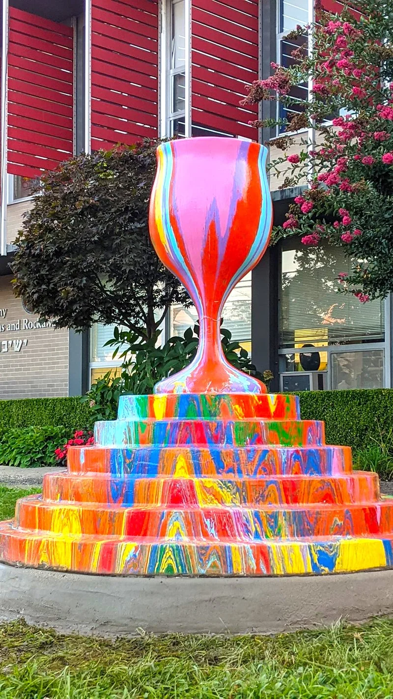 Colorful abstract sculpture resembling a large, vibrant wine glass, situated outdoors in front of a red building with windows and surrounded by trees and pink flowers.