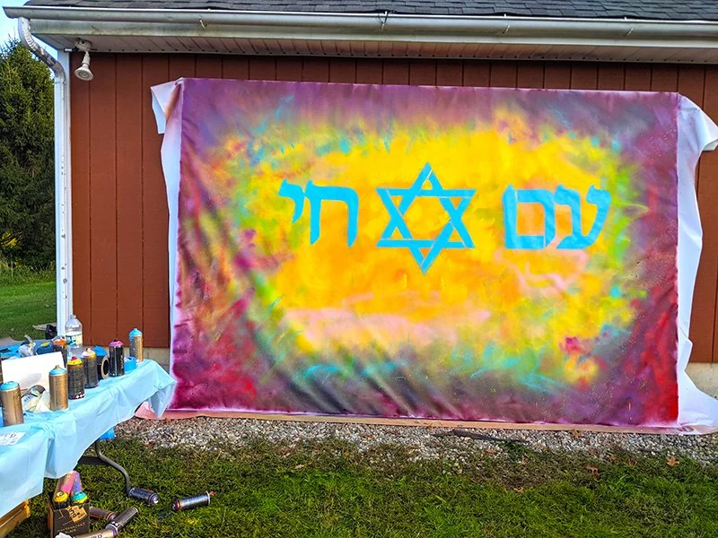 Colorful tie-dye fabric with the words 'Love' and 'Hate' in Hebrew in blue, a Star of David in the middle, hanging on a wall outside.