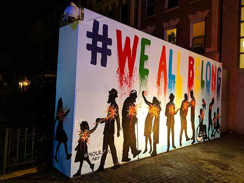 Colorful outdoor mural with silhouettes of people and a bicycle, and the hashtag '#WEALLBEHONG' written in rainbow colors.