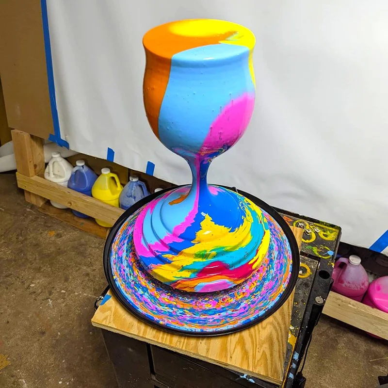 Colorful swirl pattern created with paint poured into a wine glass and onto a large plate, with bottles of paint and supplies in the background.