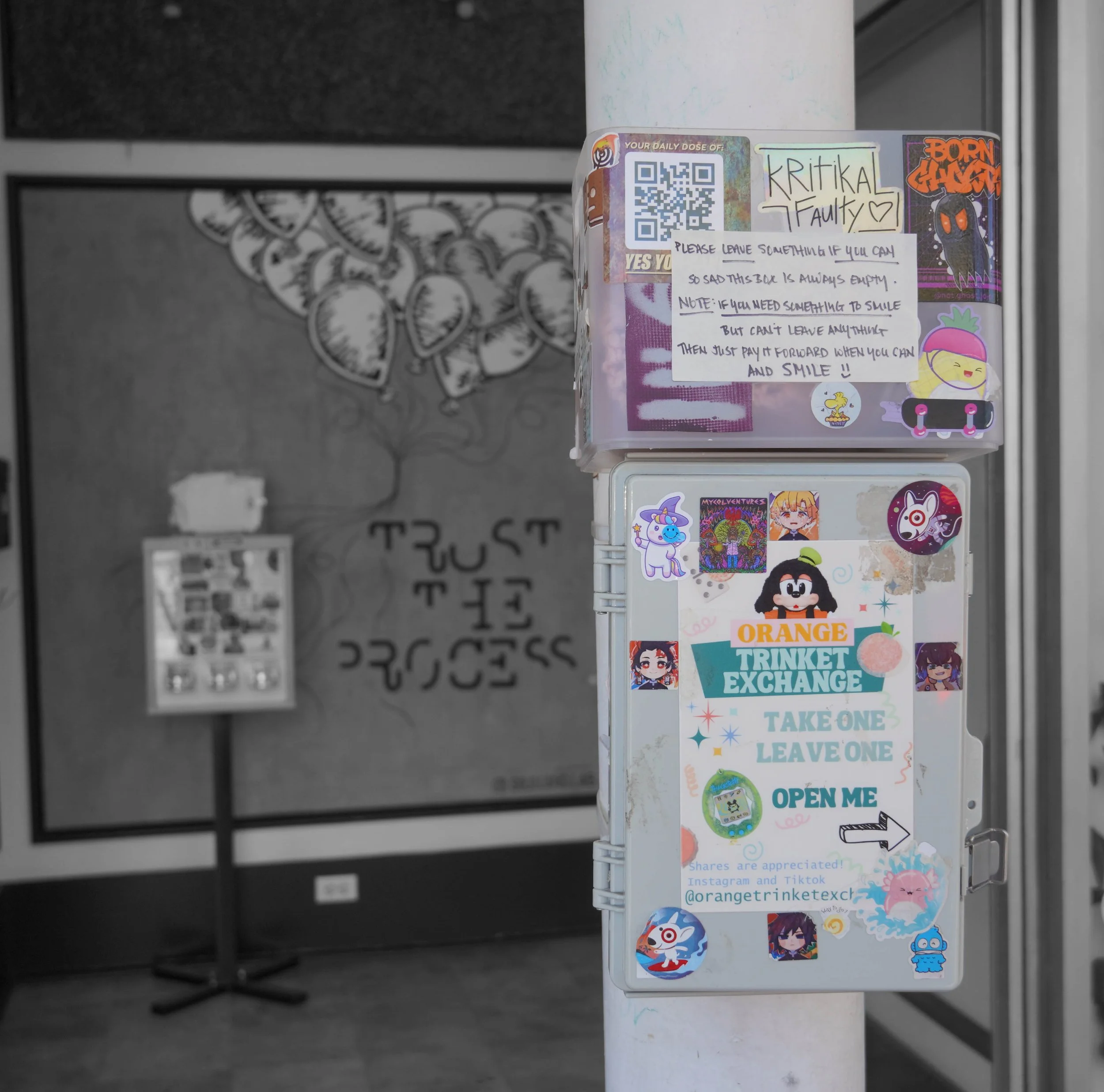Photo of a gray box attached to a vertical post adorned in version stickers and a sign that says "Orange Trinked Exchange, Take One Leave One, Open me" with an arrow to the handle.