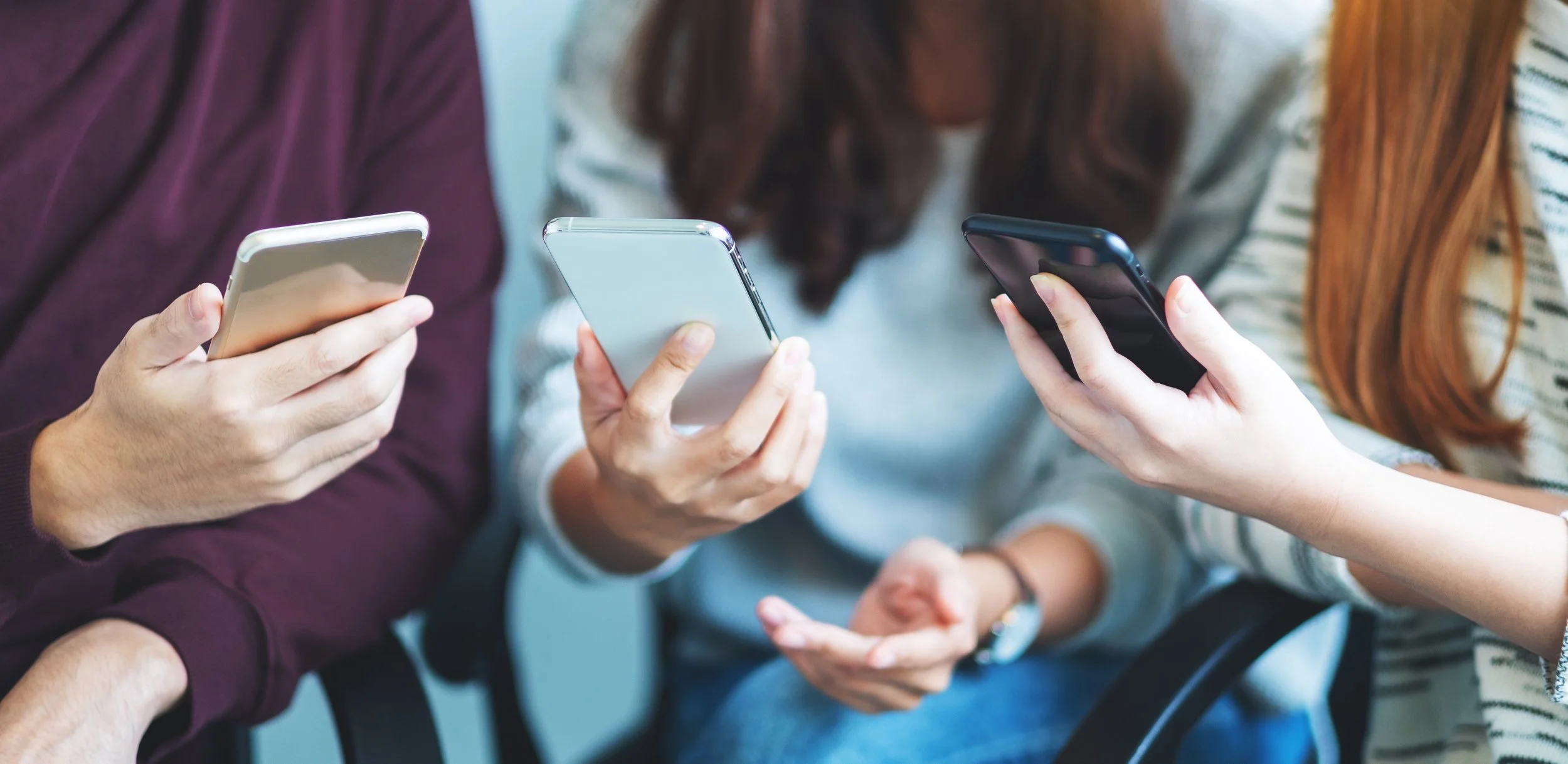 Three people sit side by side, each looking at a smartphone; their focused posture suggests quiet connection through technology in a casual setting.