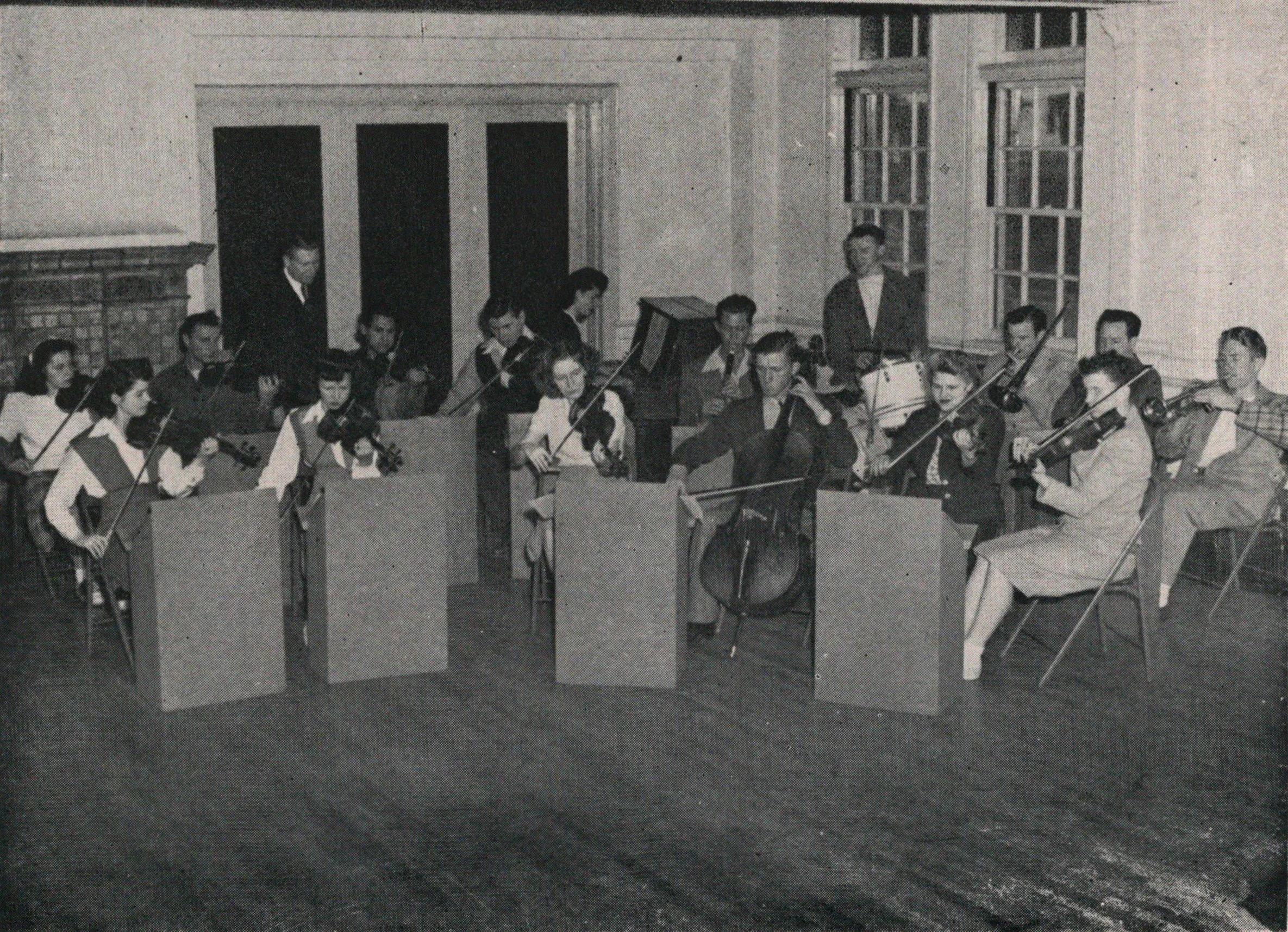 A black-and-white photo of a small orchestra rehearsing in a bare room, musicians seated behind music stands with violins and a cello, in a focused, formal mood.