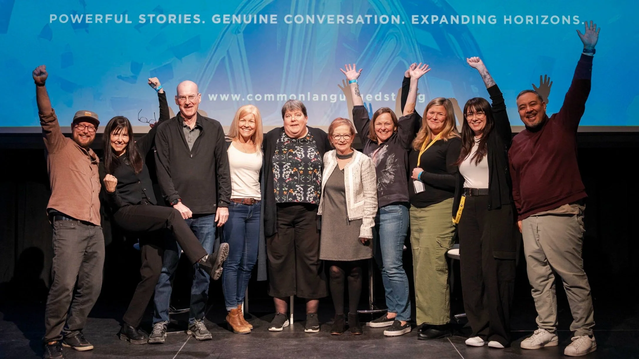 A group of eleven diverse people standing on stage with arms raised in celebration, in front of a blue background with text about storytelling and conversation.