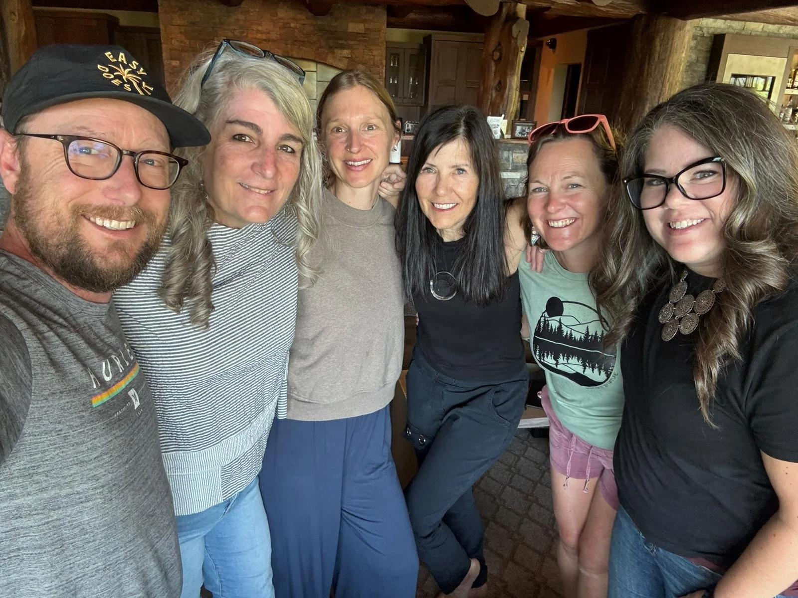 Six people standing close together indoors, smiling at the camera, with a rustic wooden background.