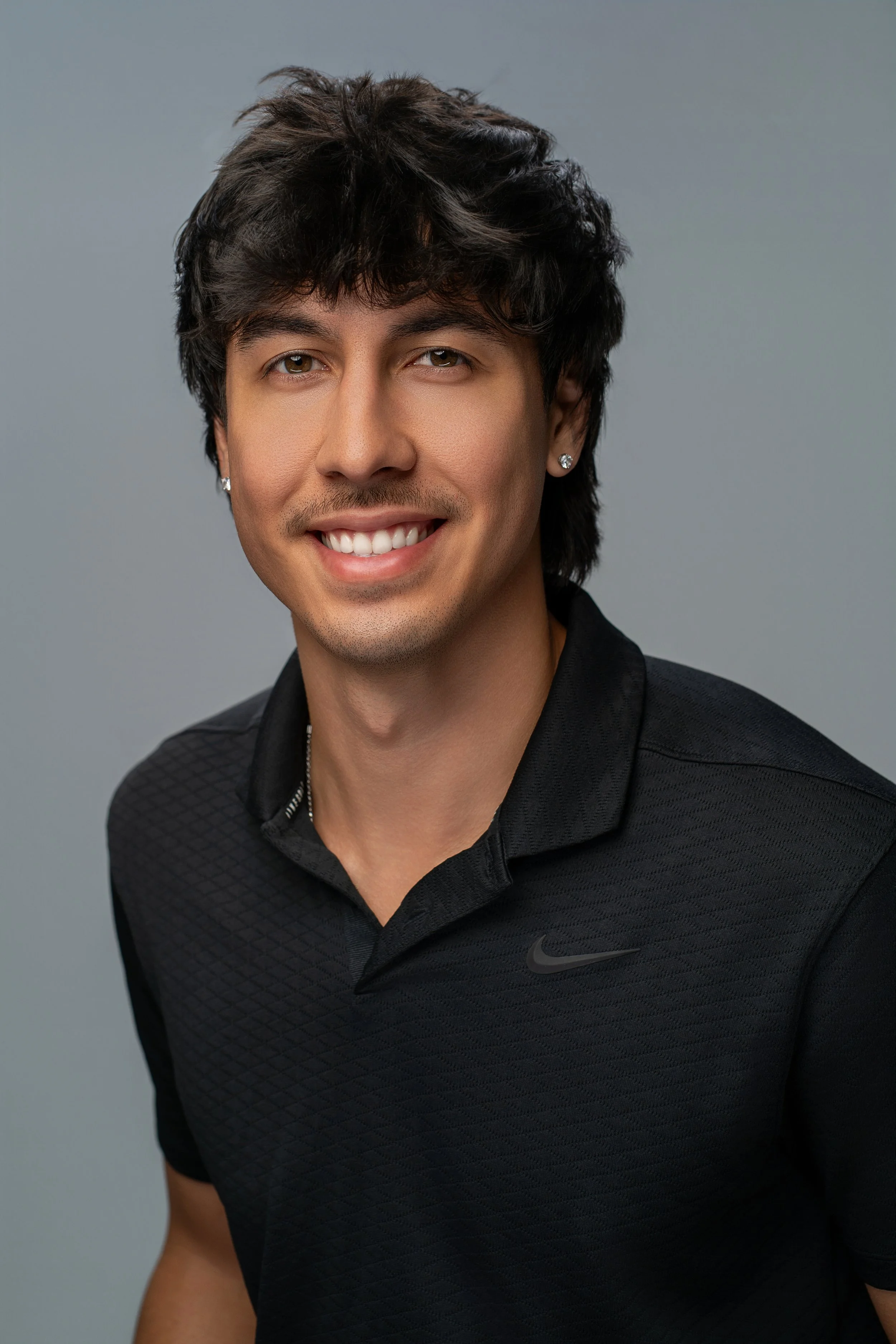 A young man with dark, wavy hair and light brown skin, smiling, wearing earrings and a black Nike shirt with a textured pattern.