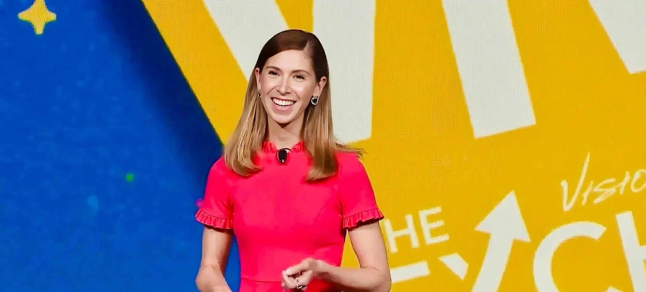Kristel Bauer with shoulder-length hair, wearing a bright pink dress, speaking on stage with a yellow and blue background.