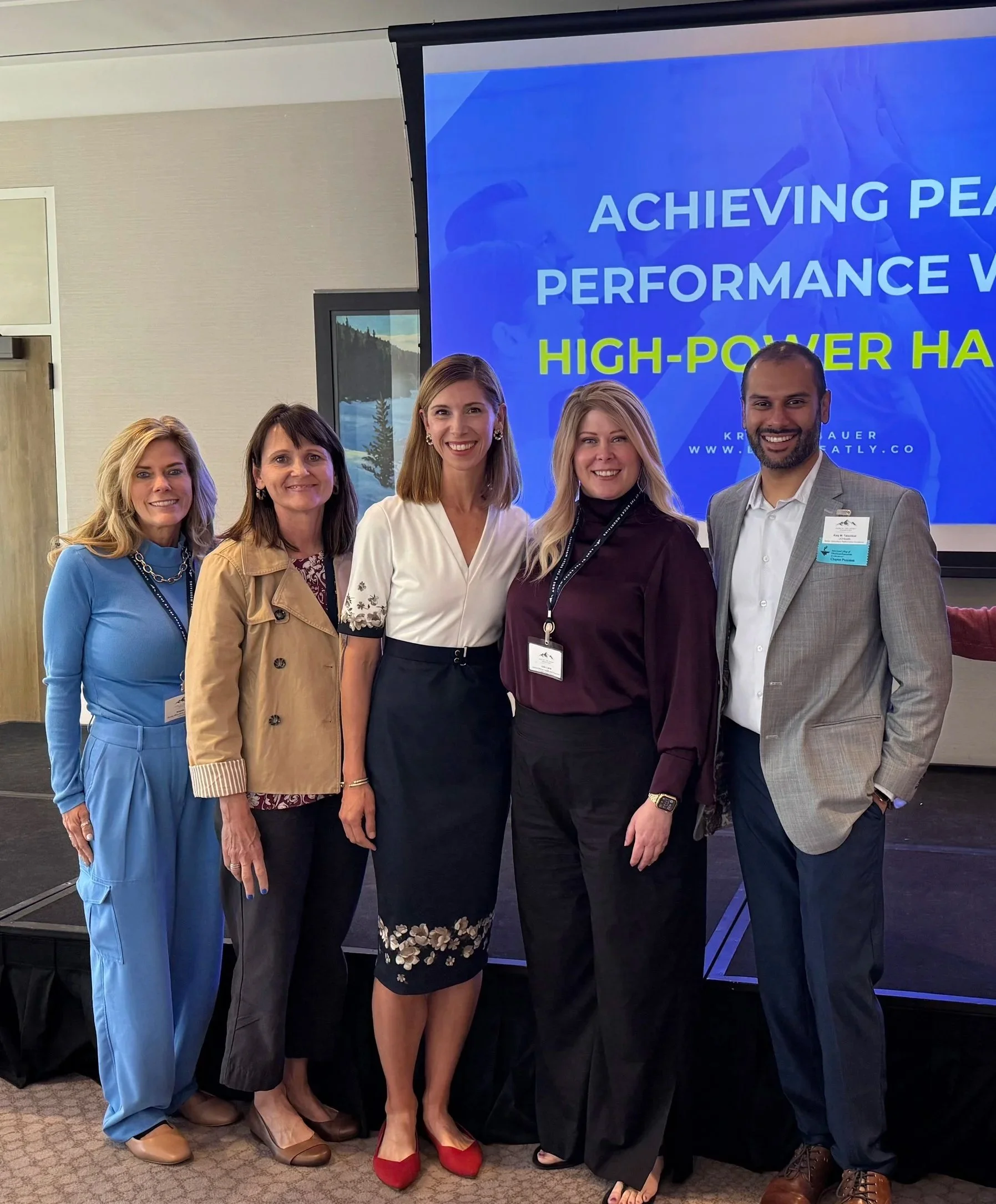 Five people standing together in front of a conference presentation screen, smiling, with a blue slide displaying 'Achieving Peak Performance with High-Power Habits.'