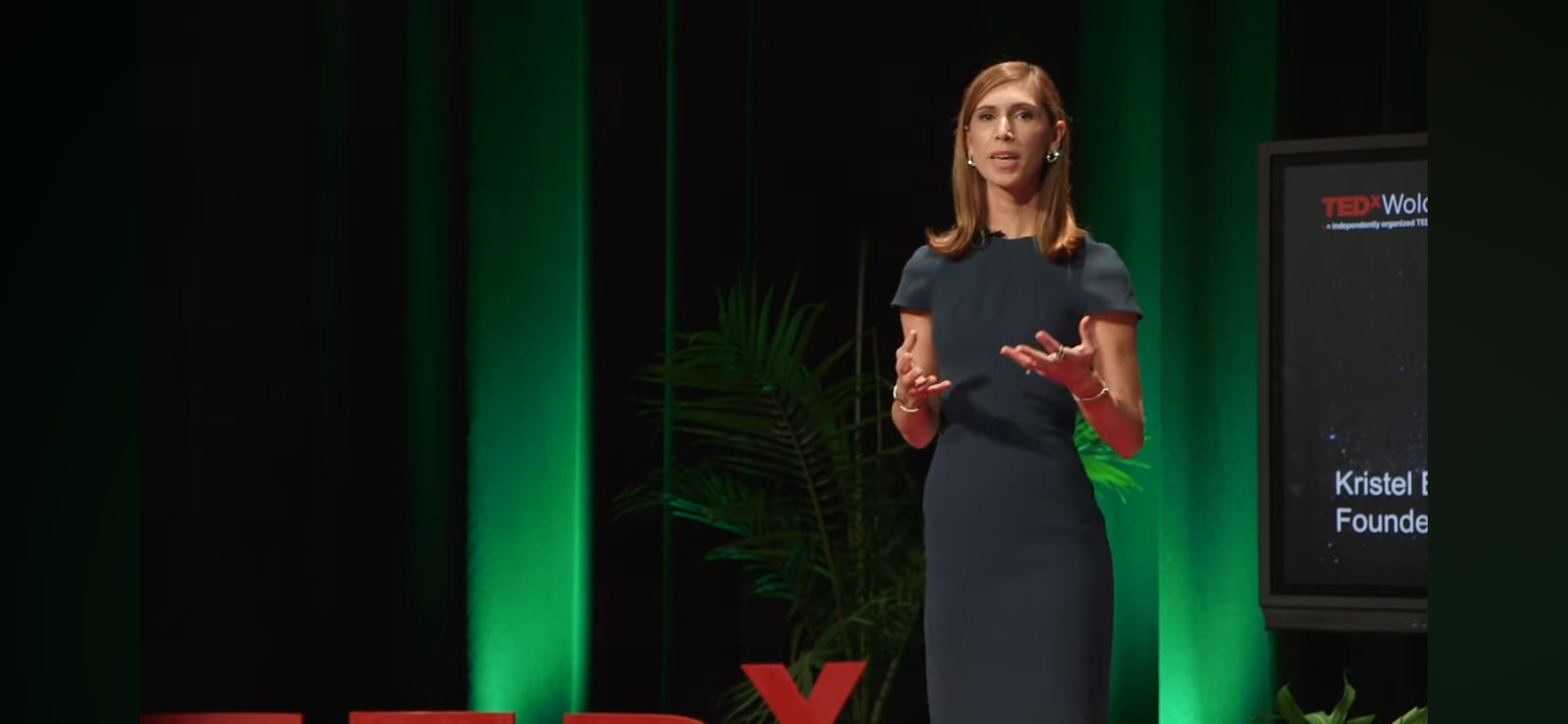 Kristel Bauer in a dark dress giving a presentation on stage at a TEDx event, with green and black backdrop and a screen displaying her name and title.