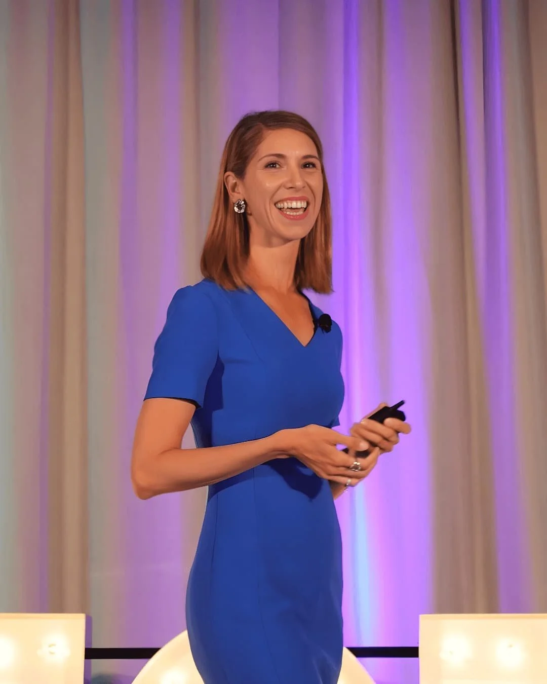 A woman in a blue dress standing on a stage, smiling, holding a remote or microphone, with a curtain background that is lit with purple lighting.