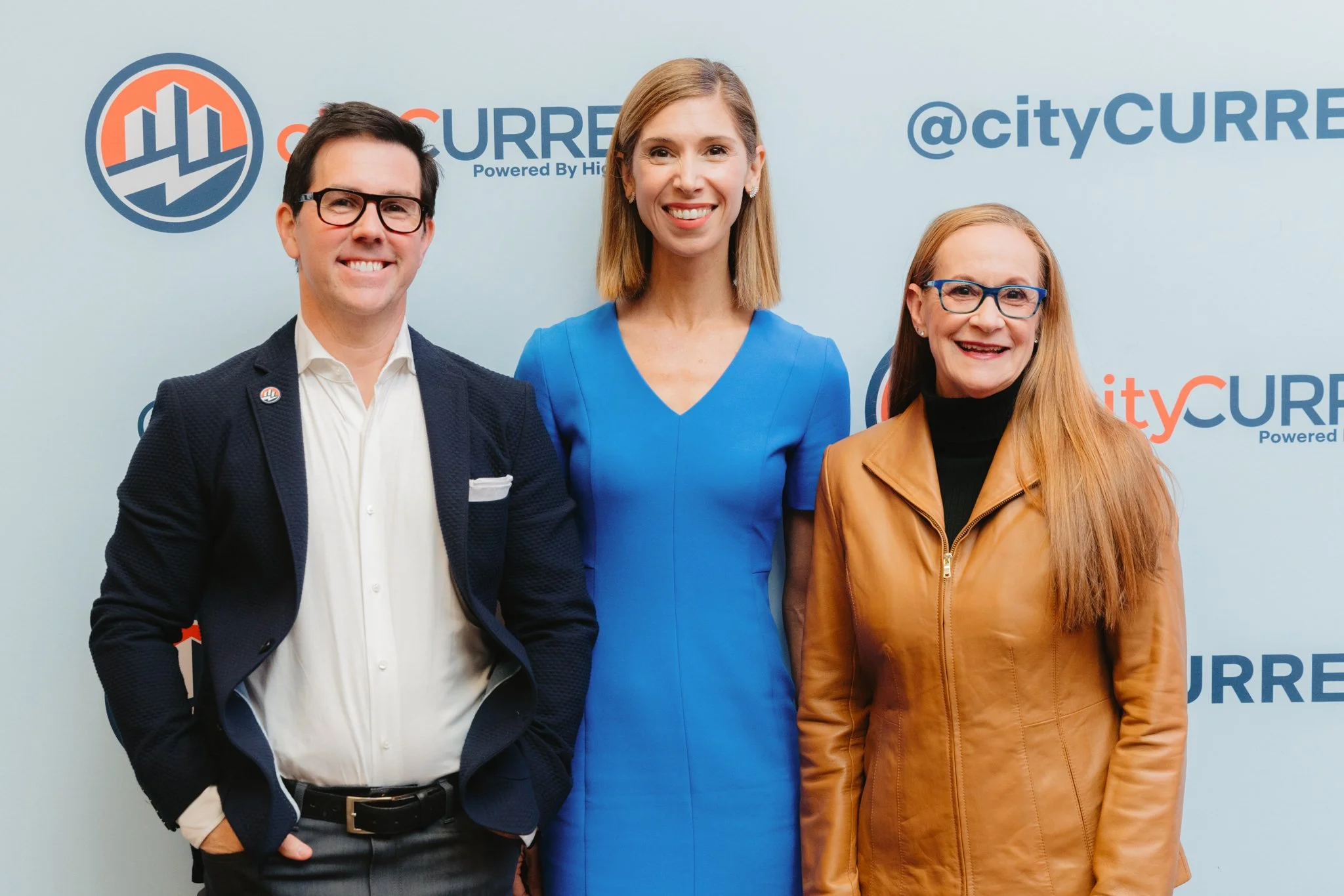Three professionally dressed people standing together and smiling at the camera in front of a backdrop with 'CityCurrent' branding.