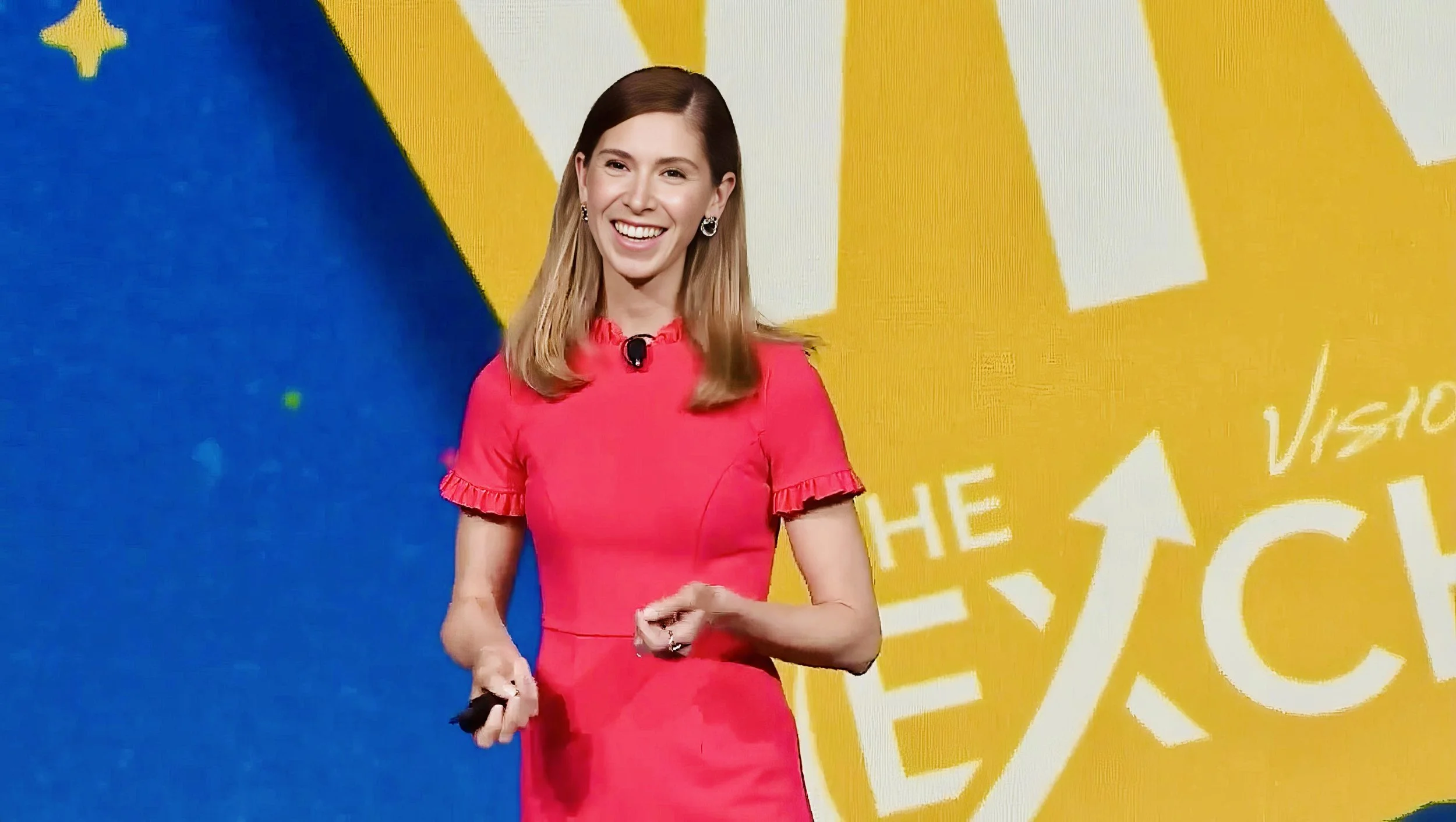 A woman smiling and holding a remote control, standing in front of a colorful background with abstract text.