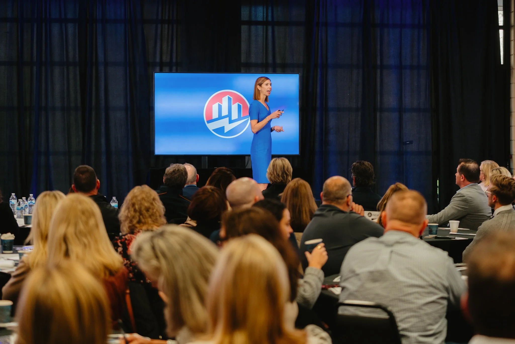 Kristel Bauer,  a woman in a blue dress giving a presentation at an event, with a large screen behind her displaying a logo with blue and red colors. The audience is seated at round tables, attentively listening.