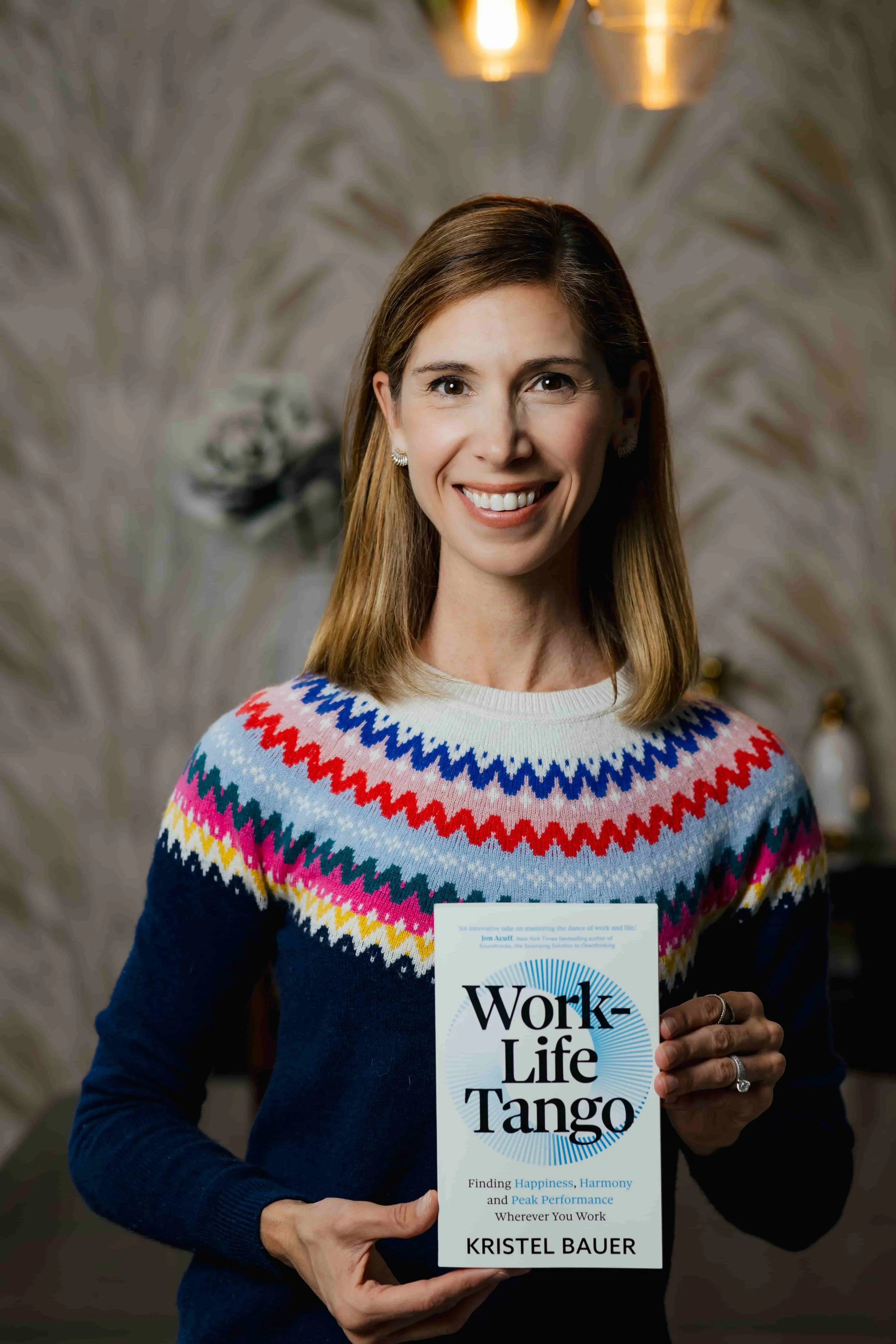 Kristel Bauer with shoulder-length brown hair smiling and holding a book titled 'Work-Life Tango' by Kristel Bauer.